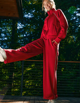 Person in a red jacket with white piping and red pants standing on a wooden deck with greenery in the background.