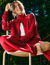 woman wearing a red zip-up jacket with white piping sitting outdoors with trees in the background.