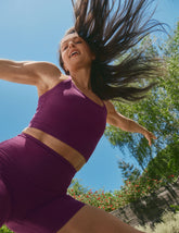 model in purple racerback cropped tank and biker shorts outdoors with trees and blue sky in the background.