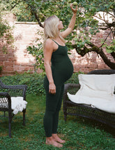woman in a green maternity midi length jumpsuit standing in a garden with a tree and chairs in the background.