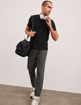 man in black t-shirt and gray joggers with pockets holding a black bag and white sneakers against a plain background.